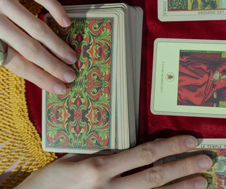 Deux mains manipulant un jeu de cartes de tarot sur une nappe rouge.