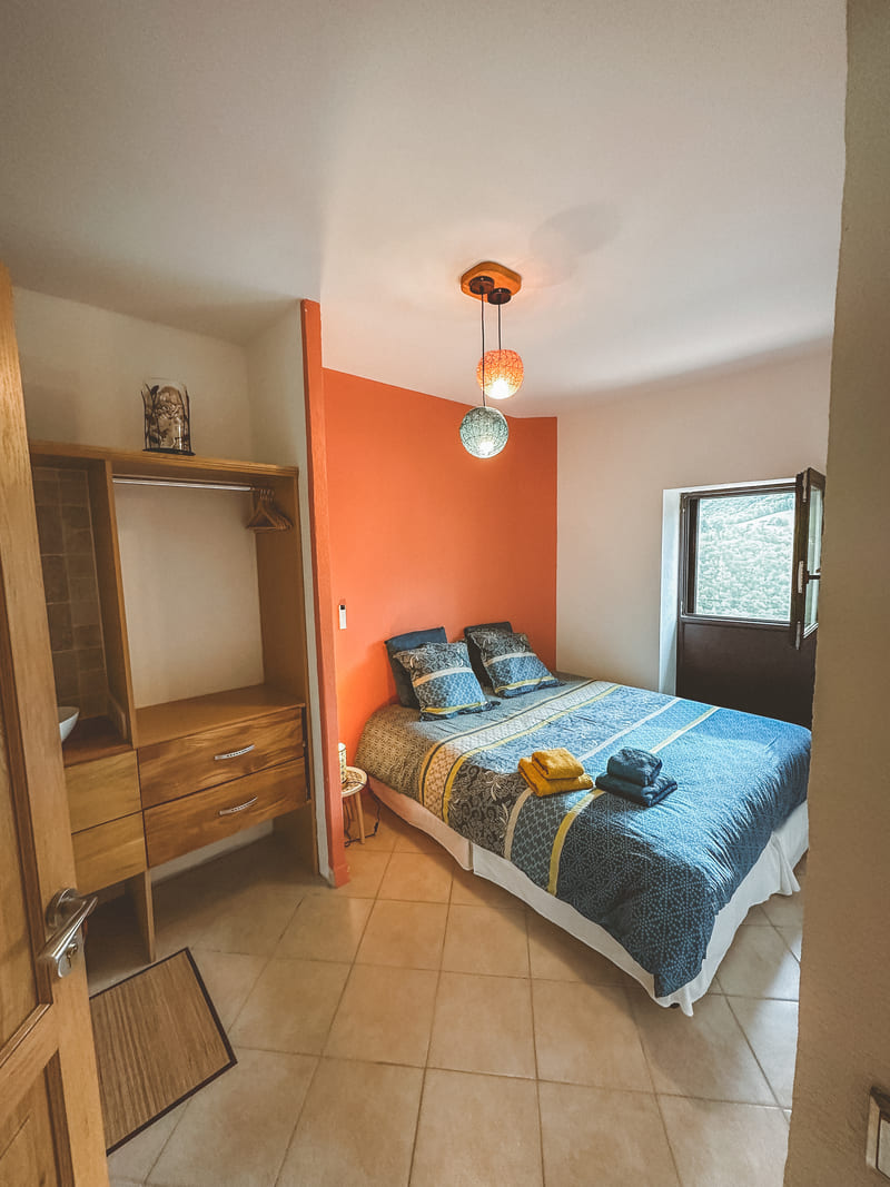 Chambre avec lit double, mur orange, étagère en bois et fenêtre ouverte.