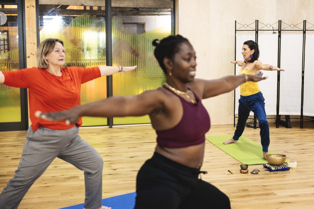 Trois femmes font du yoga dans une salle avec parquet et lumi&egrave;re naturelle.