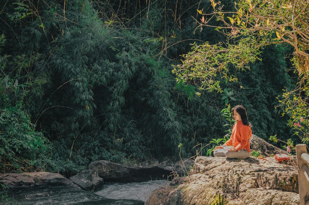 Femme m&eacute;ditant sur un rocher pr&egrave;s d'une rivi&egrave;re, entour&eacute;e de verdure.