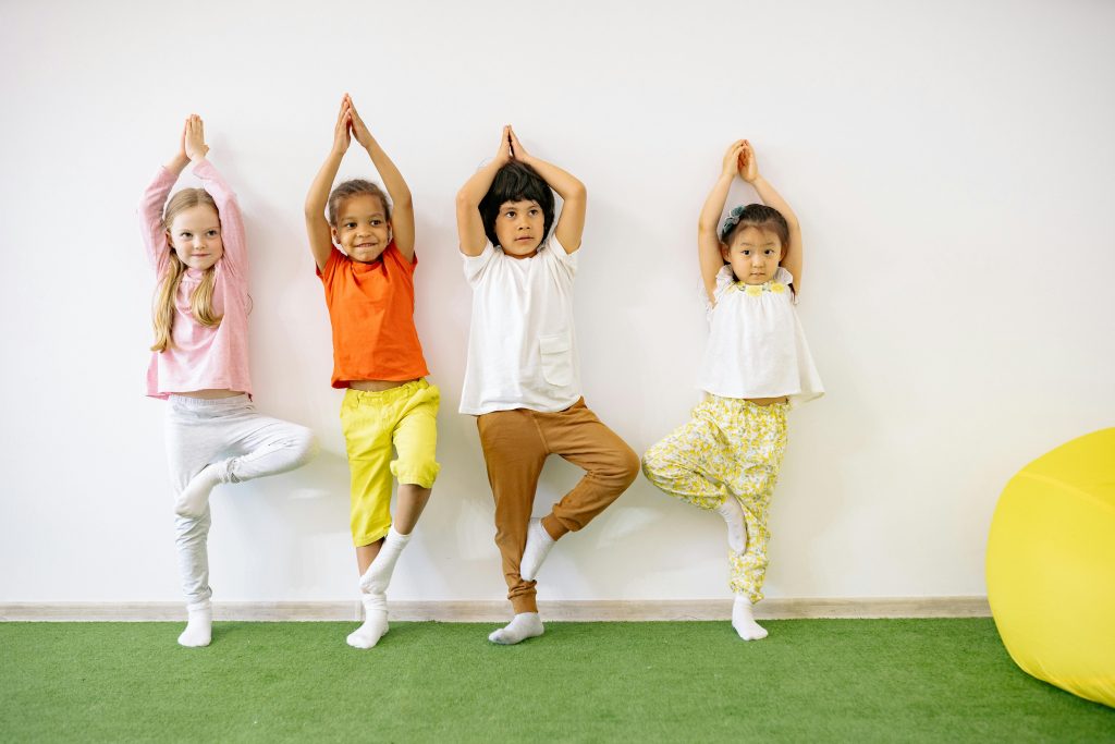 Quatre enfants font du yoga en posture de l'arbre sur un sol vert.