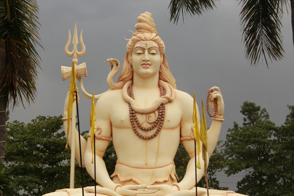 Statue de Shiva avec trident et serpent, entourée de palmiers.