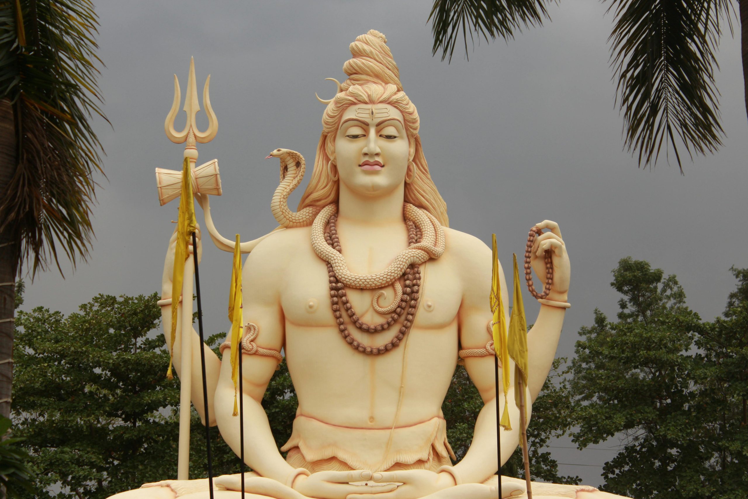 Statue de Shiva avec trident et serpent, entourée de palmiers.